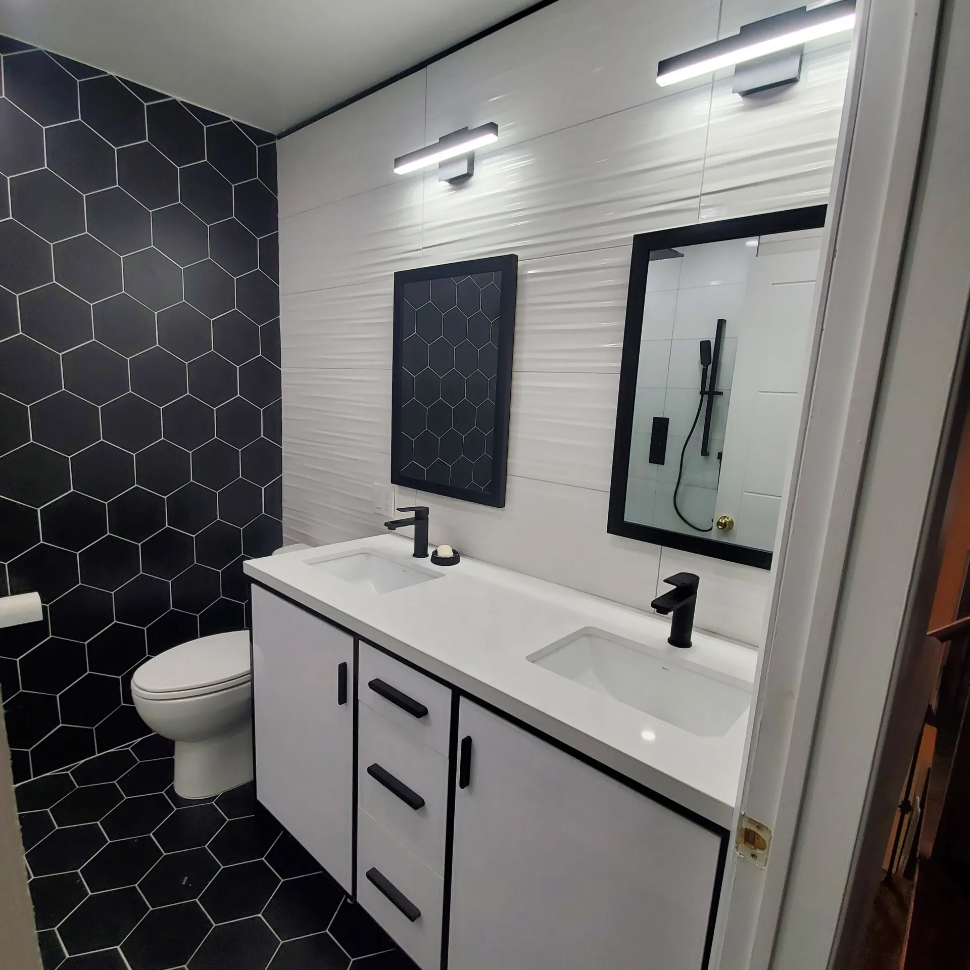 Modern bathroom with double white vanity, black hexagon tile wall, dual black faucets and mirrors