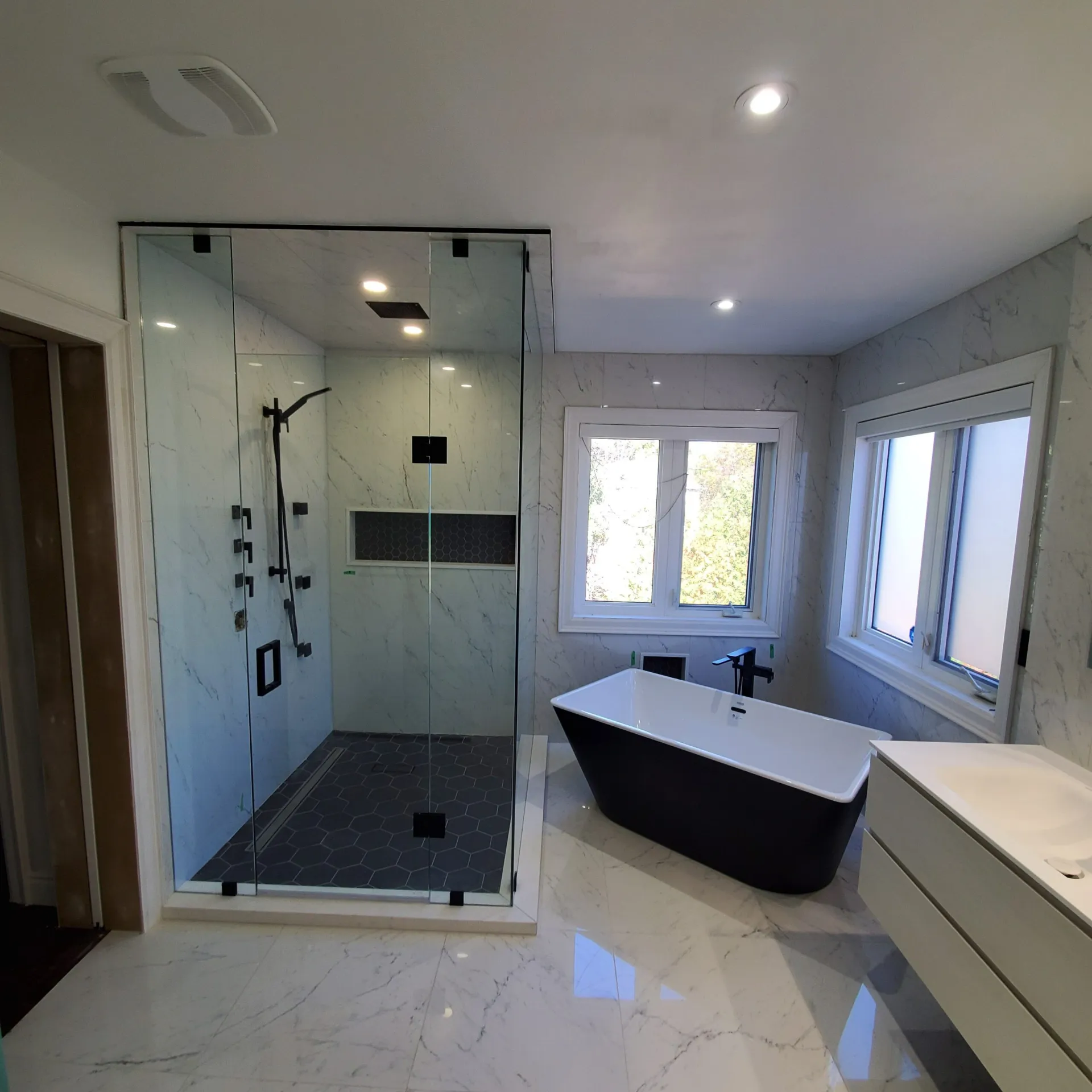 Renovated bathroom with glass shower enclosure, freestanding black soaker tub, marble tile and pot lights